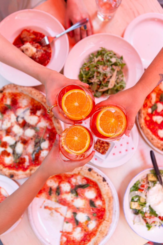 Three hands cheersing Aperol spritz cocktails over top of a table full of pizza